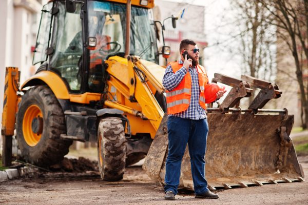 Beard worker man suit construction worker in safety orange helmet, sunglasses against tractor with mobile phone at hand.